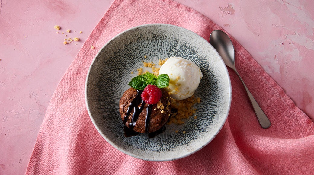 Chocolate cake with chocolate sauce, raspberry and mint leaves, plus a scoop of vanilla ice cream in a grey bowl on a pink cloth. A spoon lies beside it.