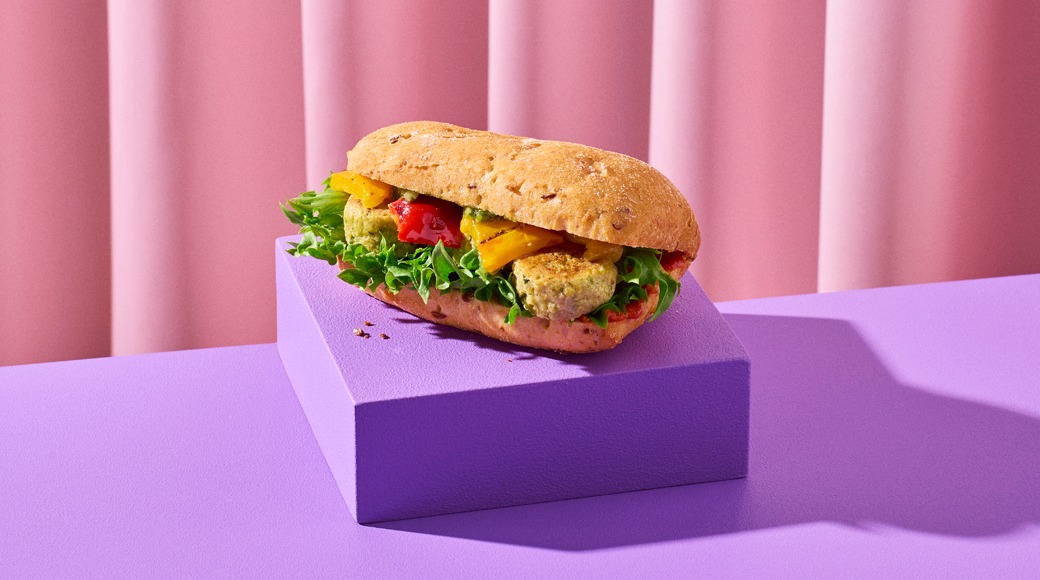 Patonki, jossa on salaattia, grillattua paprikaa ja falafelpalloja. Patonki on violetin korokkeen päällä vaaleanpunaista verhoa vasten.