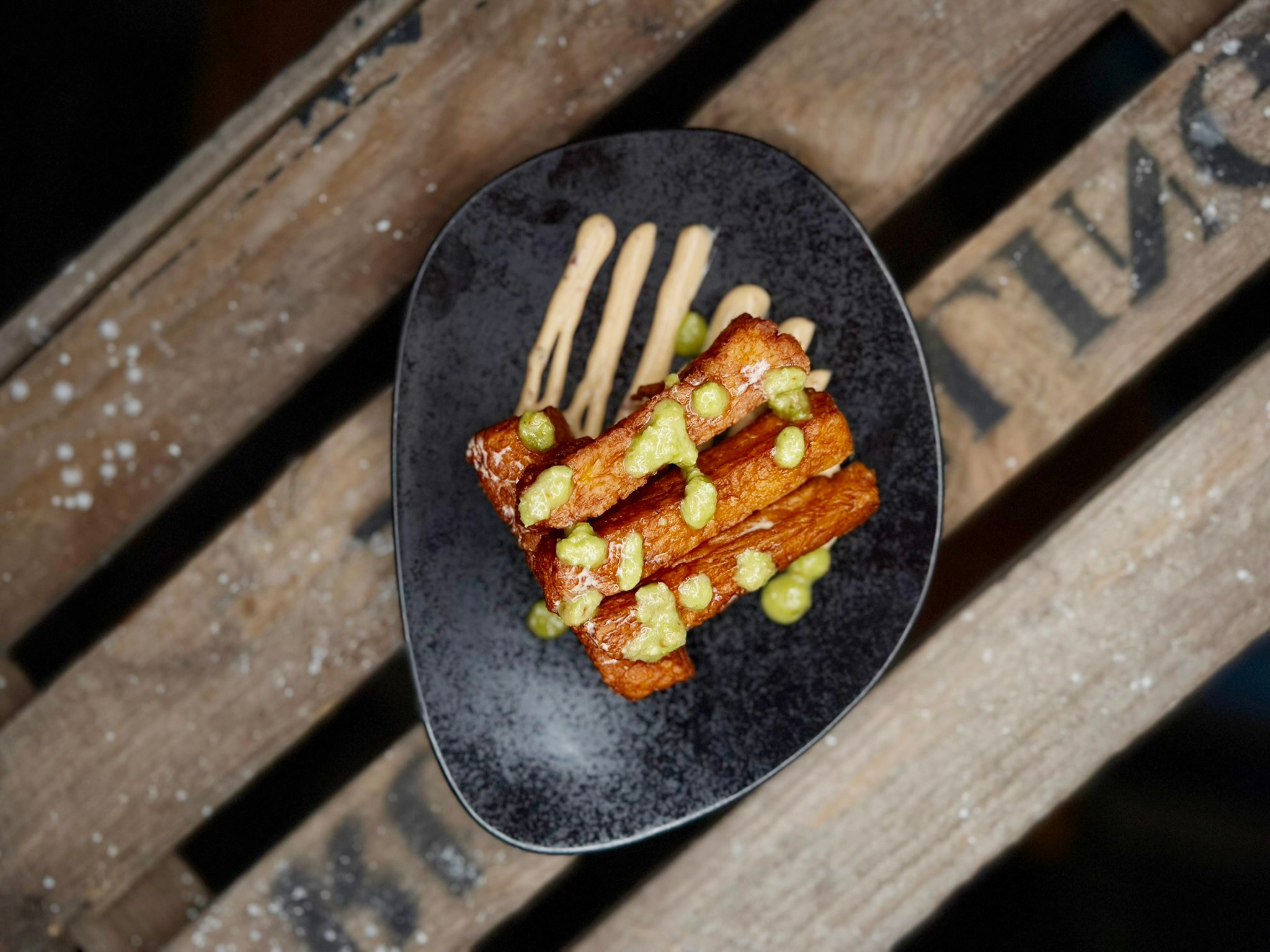 Fried, crispy brown food items with green sauce on a black plate, placed on a wooden surface.