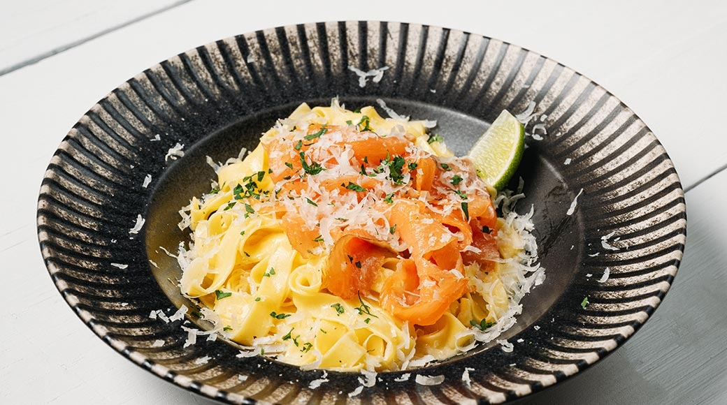 Tagliatelle suitsulõhe, riivjuustu ja värskete ürtidega tumedas vaoliste servadega kausis. Kausi serval on laimitükk.