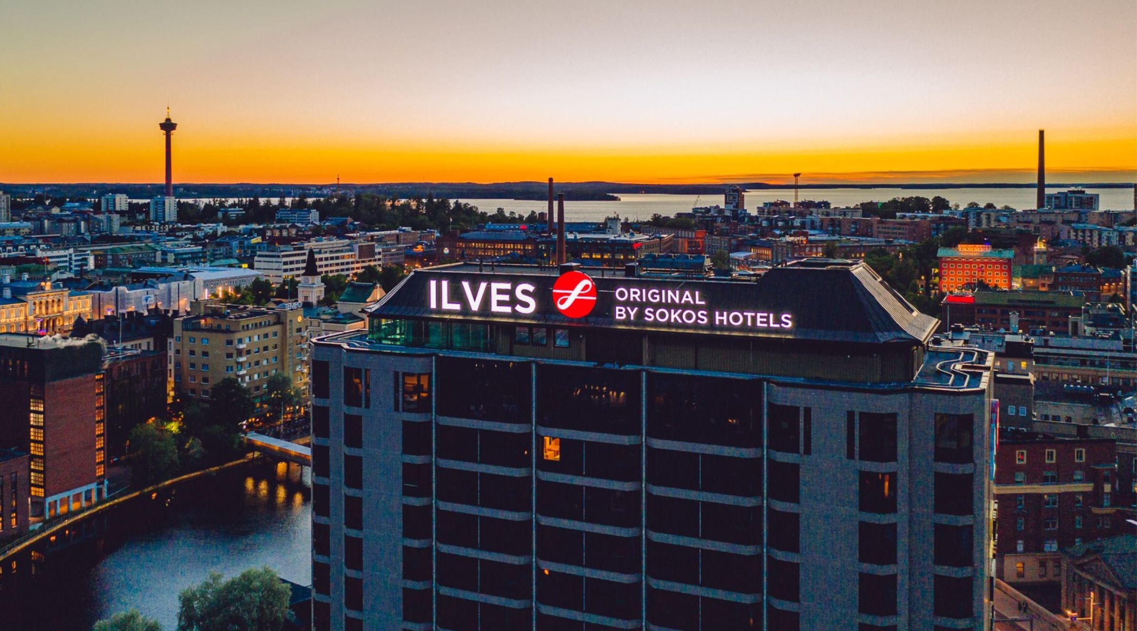 Flygfoto över Tammerfors i solnedgången med Sokos Hotel Ilves i förgrunden och Näsijärvi sjön samt stadens silhuett i bakgrunden.