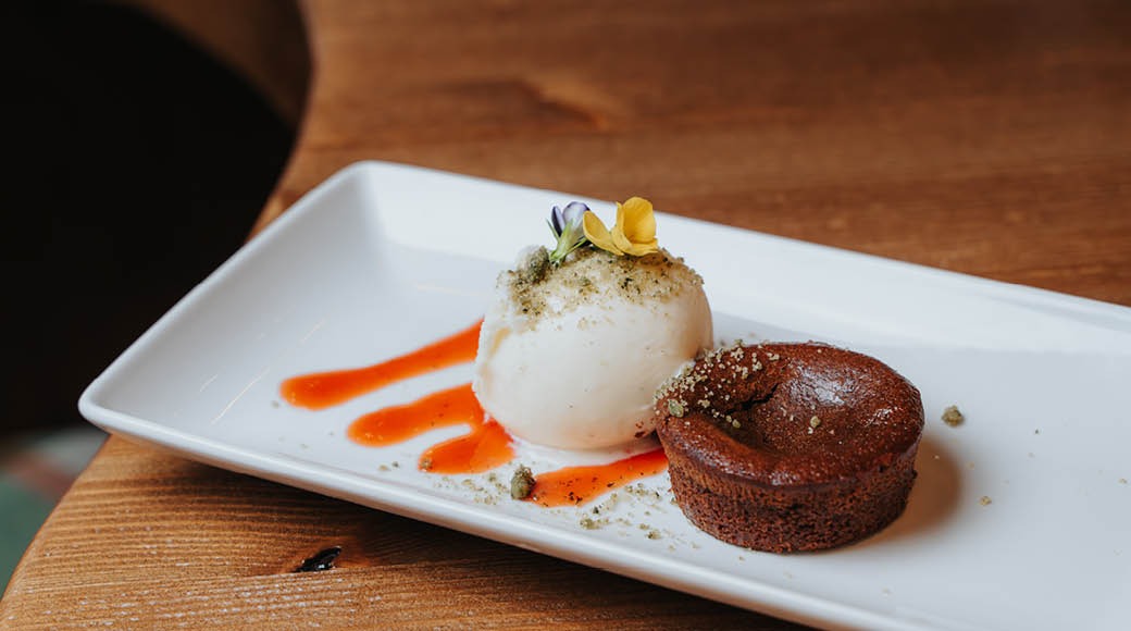 Suklaakakku ja jäätelöpallo koristeellisella valkoisella lautasella, jossa oranssia kastiketta puisella pöydällä.