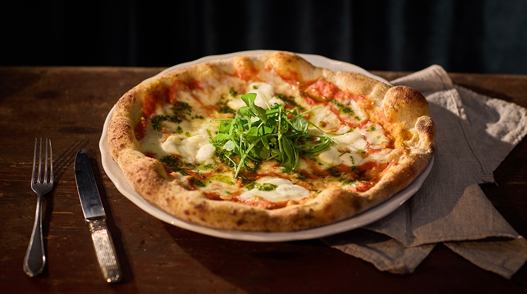 Pizza på vit tallrik, toppad med färsk rucola, röd tomatsås och mozzarella. Gaffel och kniv bredvid på träbord.