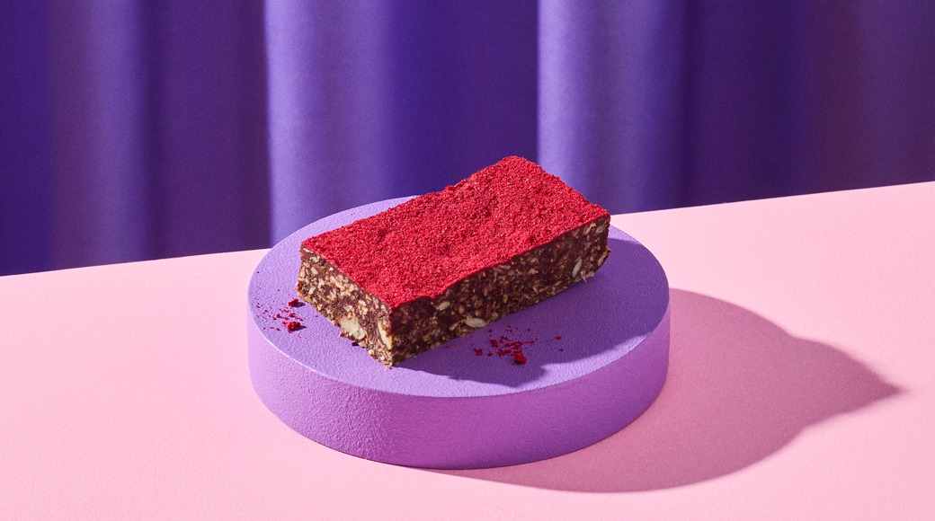 Raakapatukka, jonka päällä on punaista vadelmajauhetta, violetilla korokkeella vaaleanpunaisella pöydällä. Taustalla violetti verho.