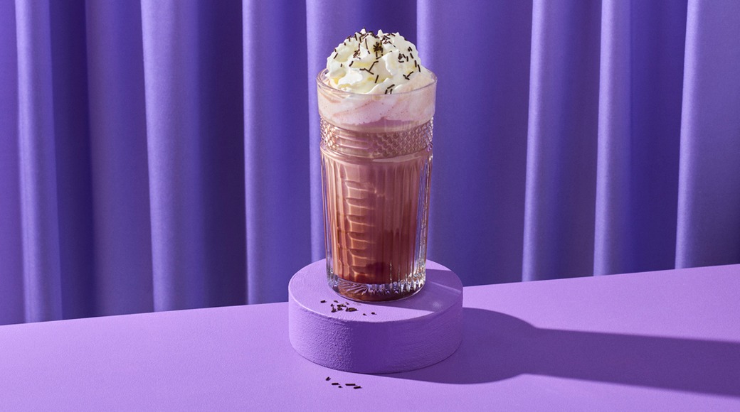 Kuviolasillinen kuumaa suklaata, päällä kermavaahtoa ja suklaarouhetta. Lasi on violetilla korokkeella violettia verhoa vasten.