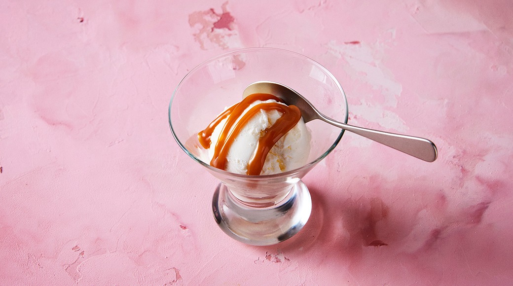 Scoop of vanilla ice cream with caramel sauce in a glass dish with a spoon. The dish is on a pink surface.