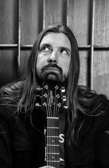 Black and white portrait of a long-haired, bearded man looking upward behind the neck of an acoustic guitar. Wooden panelling in the background.