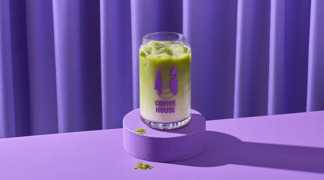 Jäinen matcha latte lasissa violetin korokkeen päällä violettia verhoa vasten. Matchajauhetta pöydällä.