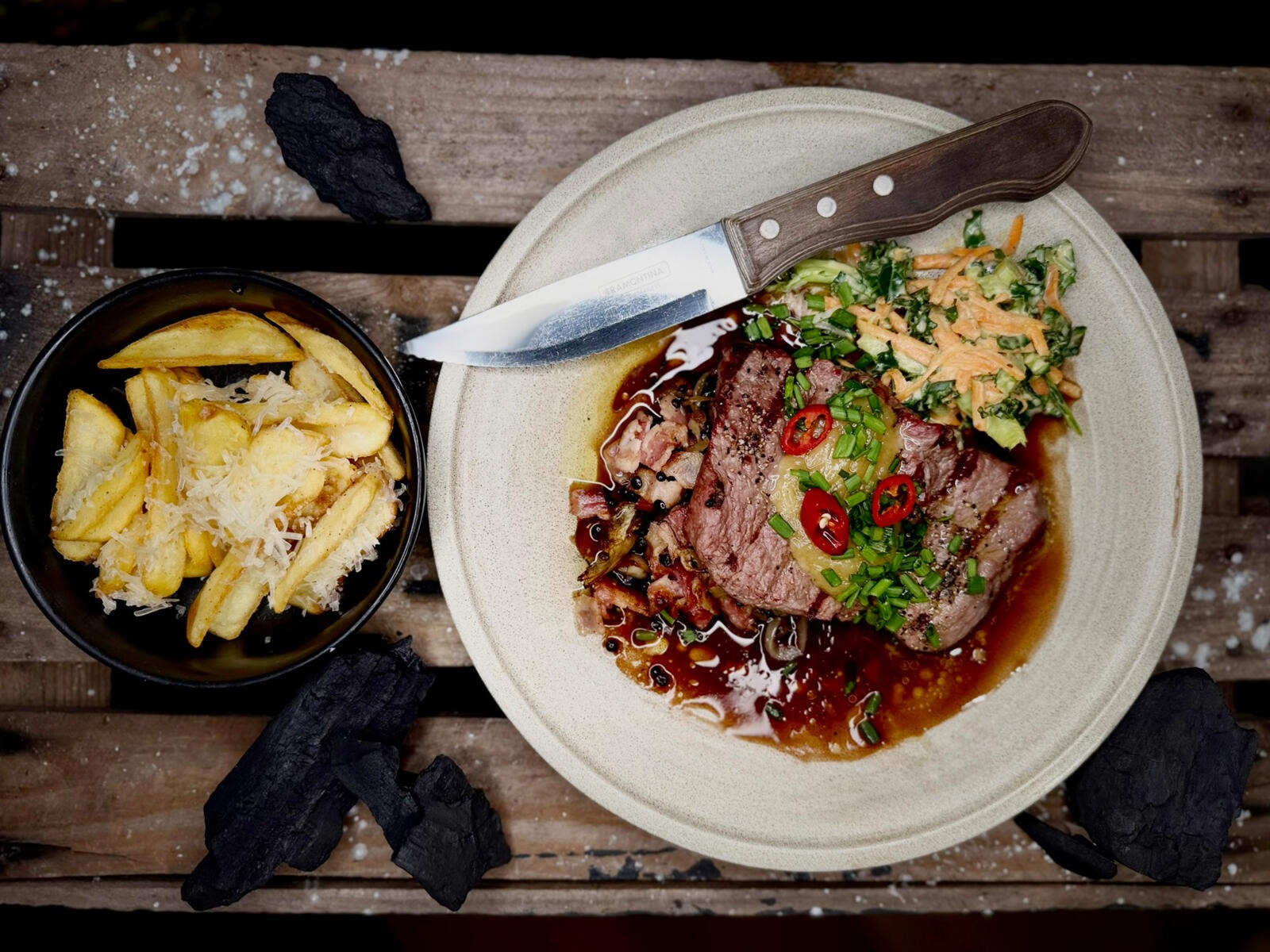 A plate of beef in sauce garnished with chilis and herbs, next to cheesy fries. Served on wooden surface.