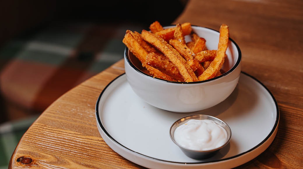 Kullanruskeat bataattiranskalaiset valkoisessa kulhossa puisella pöydällä, vieressä dippikastiketta pienessä astiassa.