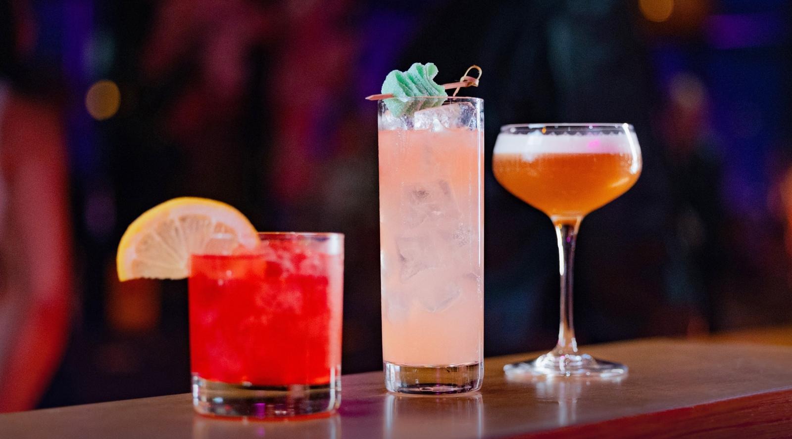 Tre olika cocktails på bardisken: en röd med citronskiva, en hög ljusrosa drink med myntagarnering och ett kupat glas med orange dryck med skum.