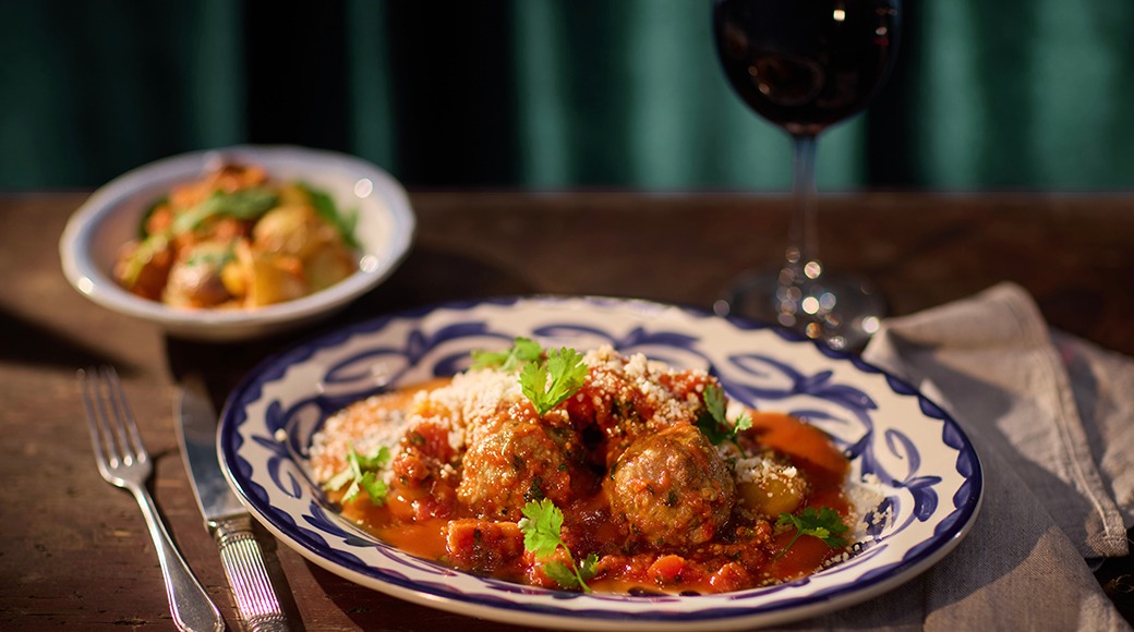 Lihapallid tomatikastmes garneeritud ürtidega, serveeritud sinise äärega taldrikul. Taustal veinipokaal ja teine toiduports.