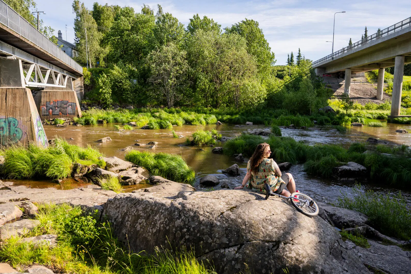 Nainen istuu selkä kameraa kohti kalliolla kosken juurella.