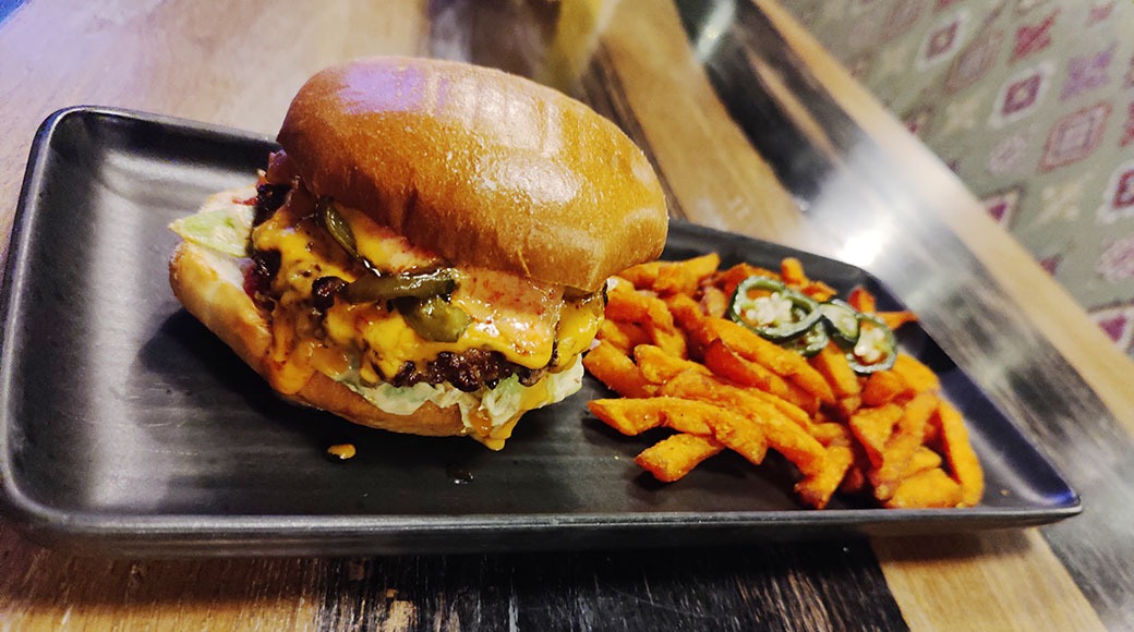 Hampurilainen ja maustetut ranskalaiset metallilavalla. Hampurilaisessa näkyy juustoa, pihviä ja salaattia pehmeän sämpylän välissä.