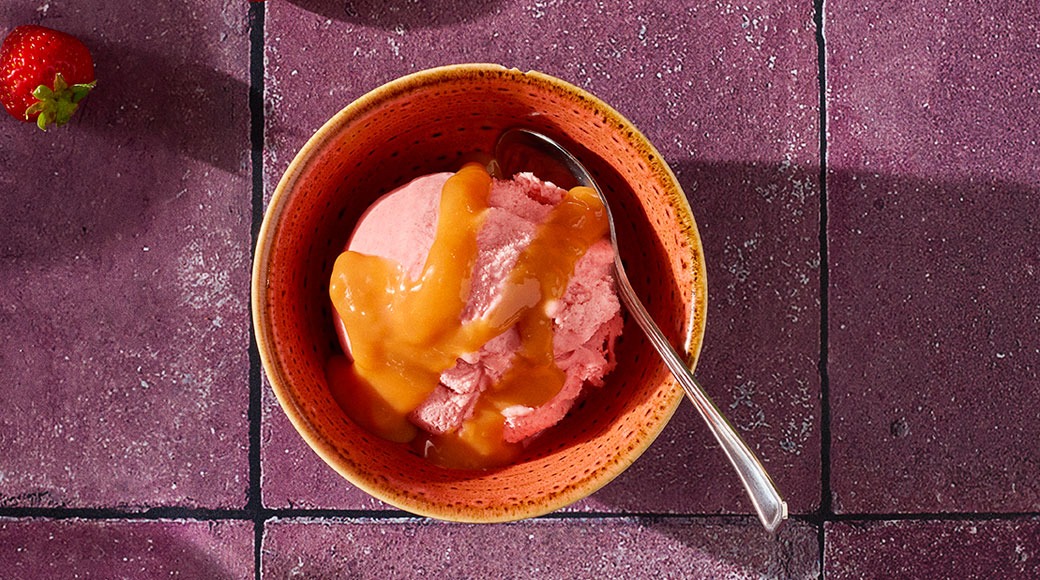 Oranssi kulho, jossa vaaleanpunaista jäätelöä ja karamellikastiketta. Kulhossa on lusikka, vieressä mansikka violetilla laatalla.