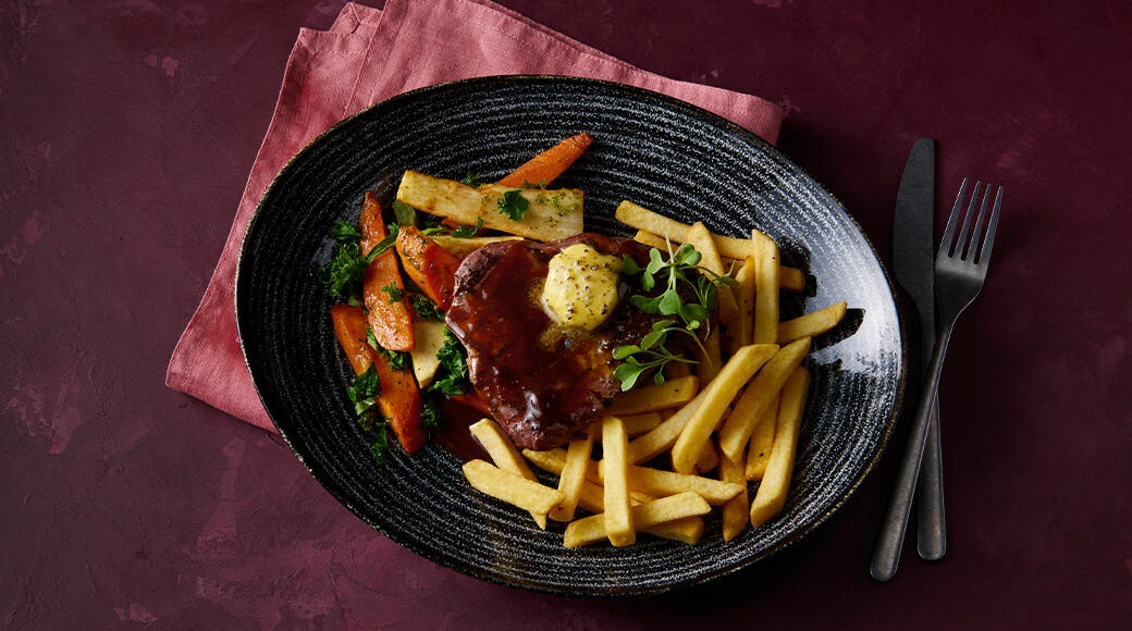 Dark plate with steak in gravy topped with a pat of butter, french fries, and roasted carrots and parsnips.