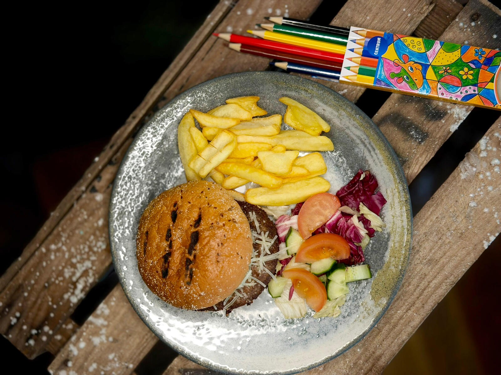 Plate with hamburger, french fries and salad on wooden surface, next to colorful pencils.