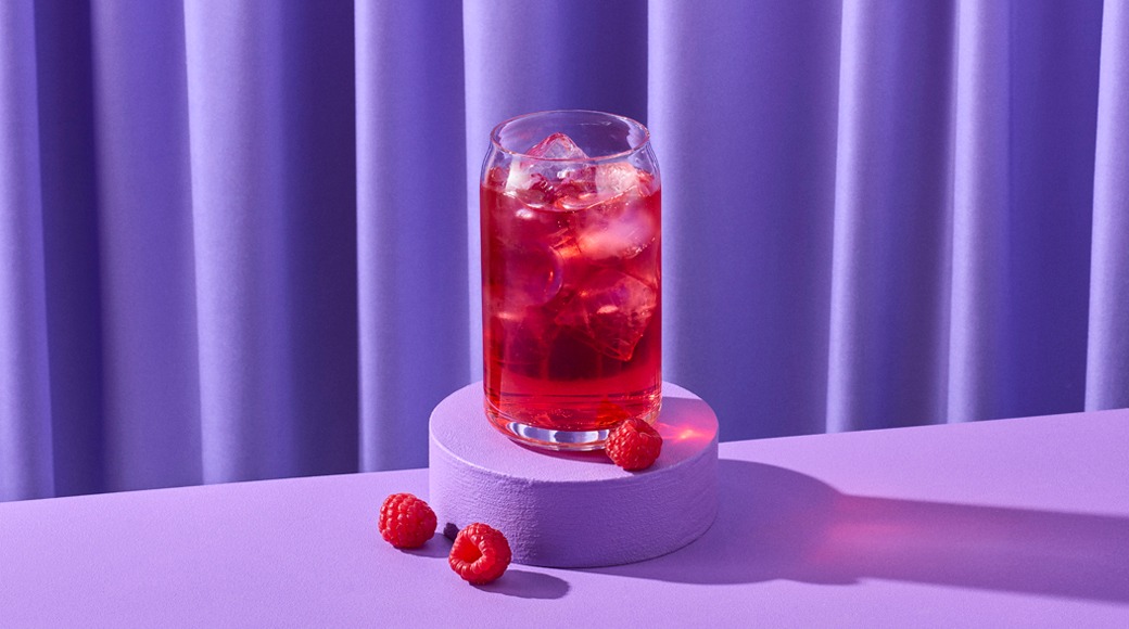 Punainen jääkylmä juoma lasissa violetin korokkeen päällä. Vieressä vadelmia, taustalla violetti verho.