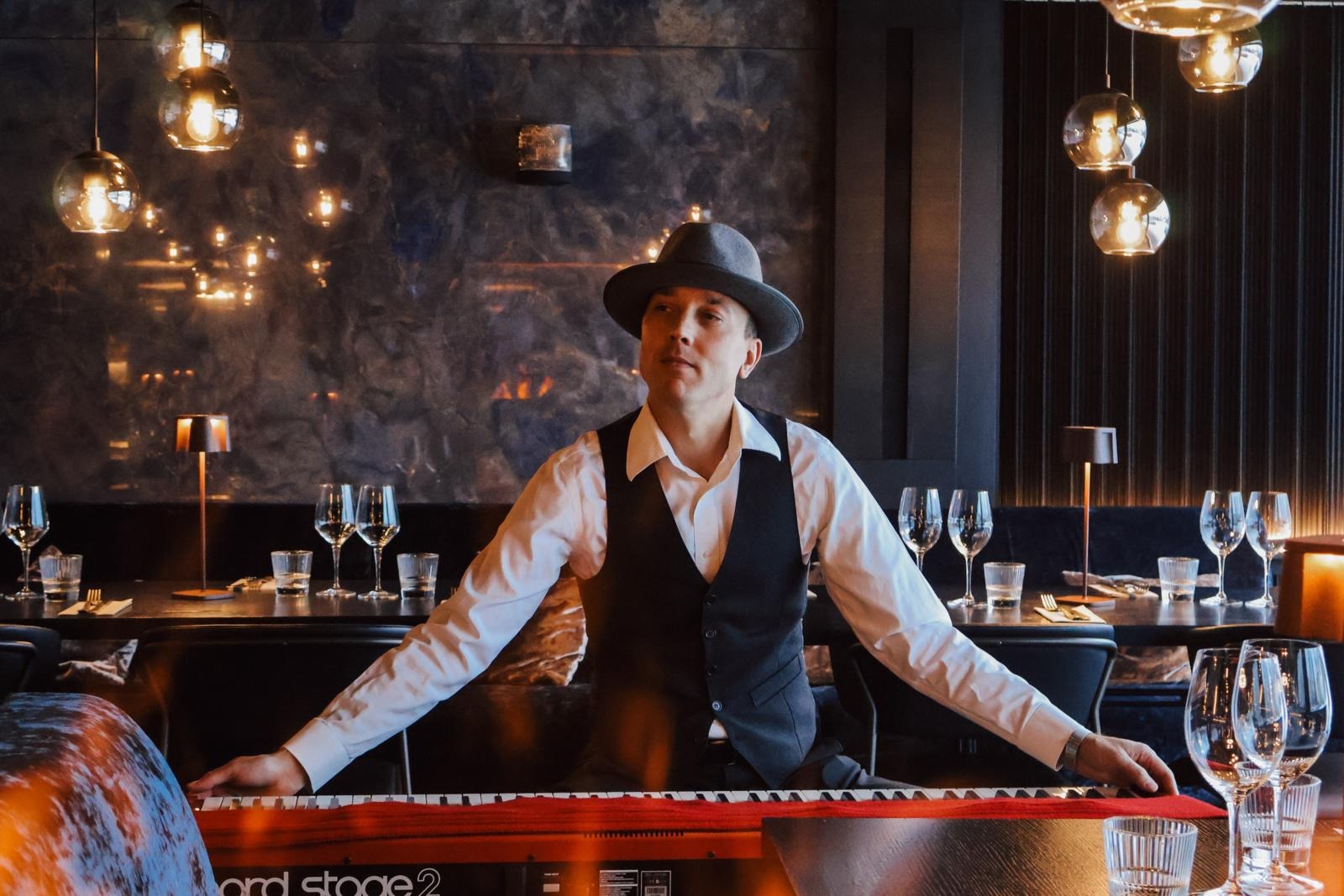 Tyylikkäästi pukeutunut pianisti soittaa ravintolan tunnelmallisessa sisustuksessa. Tilassa riippuvat pallolamput ja kattauksessa viinilasit.