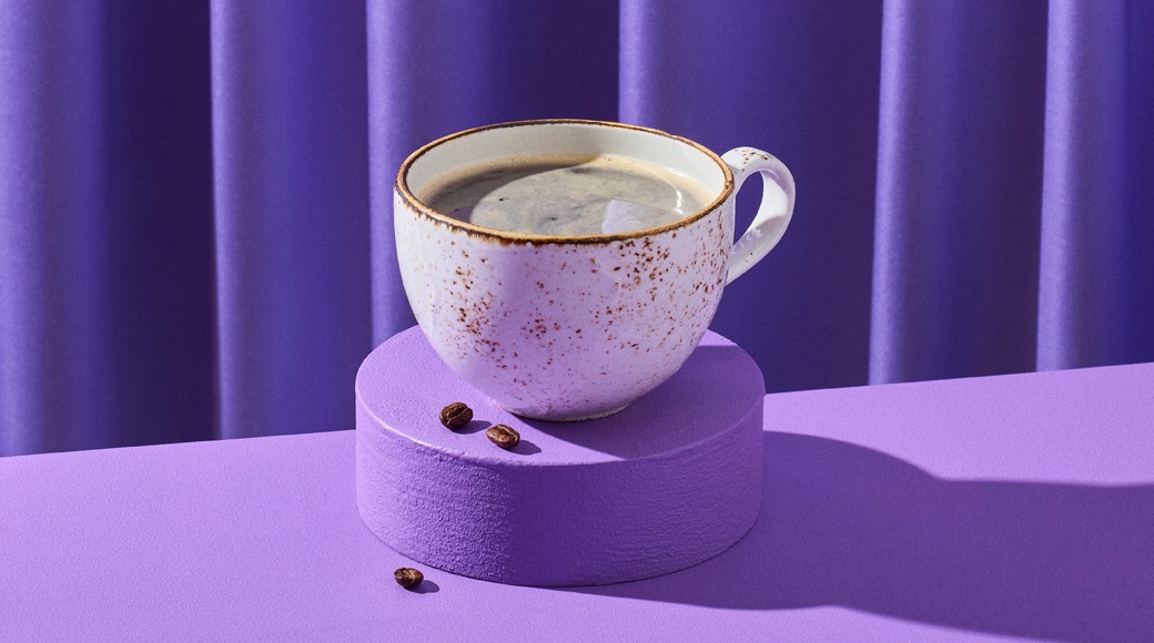 Kahvikuppi ruskeareunaisessa pilkullisessa kupissa violetin korokkeen päällä. Taustalla violetti verho, korokkeen ympärillä kahvipapuja.