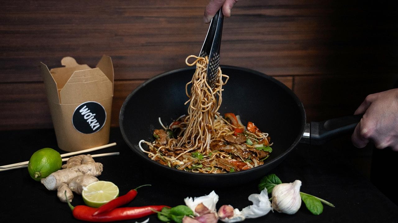 Wokkipannusta nostetaan nuudelia, taustalla takeaway-rasia.