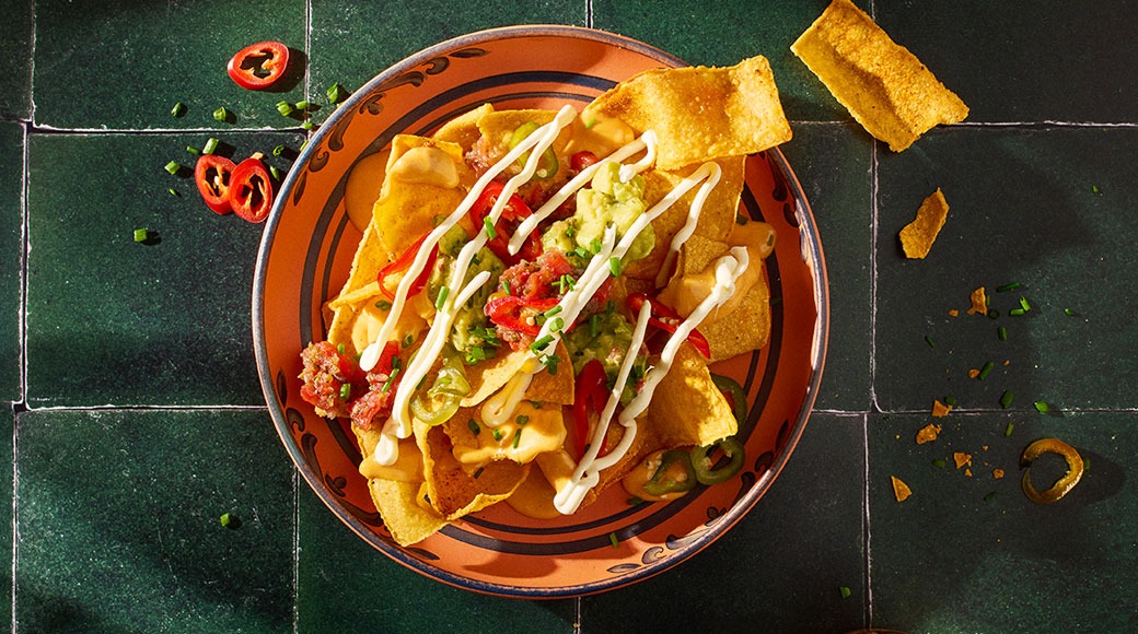 Nachoja juustokastikkeella, guacamolella, tomaattisalsalla, jalapeñoilla ja smetanasiksakilla oranssilla lautasella vihreällä laattapinnalla.