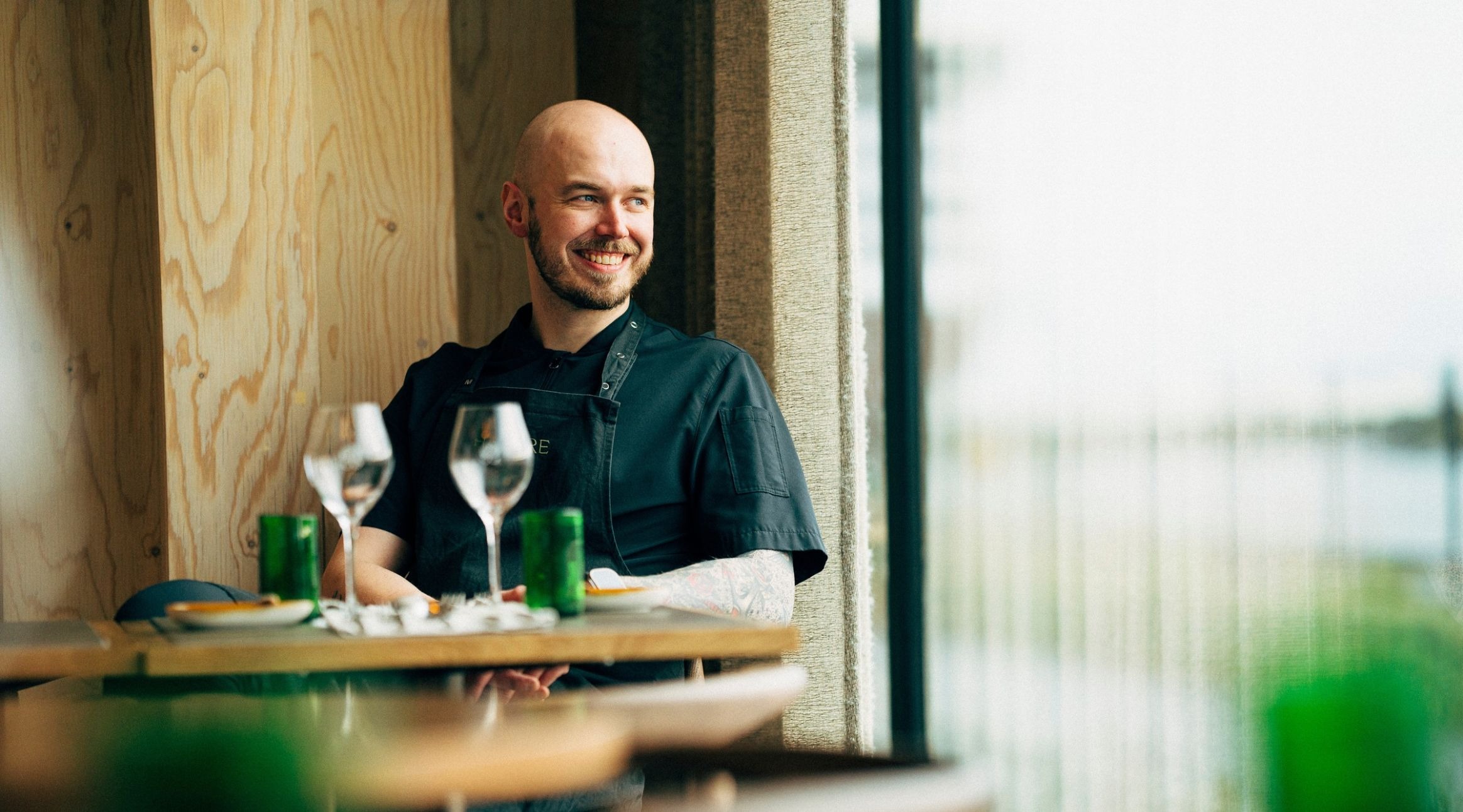 Hymyilevä kokki tummassa asussa istuu ravintolassa pöydän ääressä ja katsoo ikkunasta ulos. Pöydällä on viinilaseja ja vihreitä lasipulloja. Seinät ovat vaaleaa puuta.