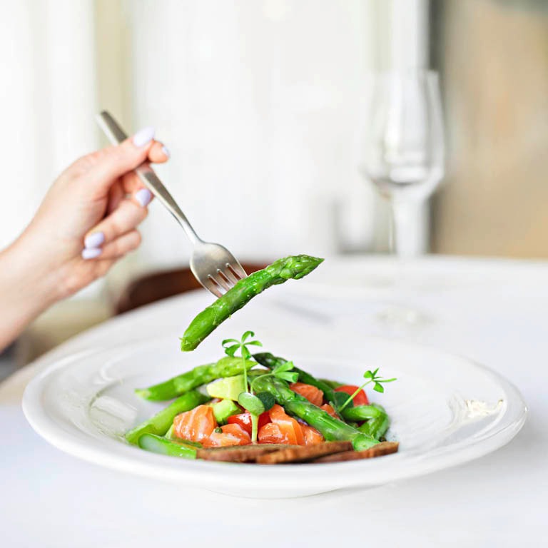 Käsi haarukkoineen nostaa parsaa valkoisella lautasella olevasta lohisalaatista, taustalla viinilasi.