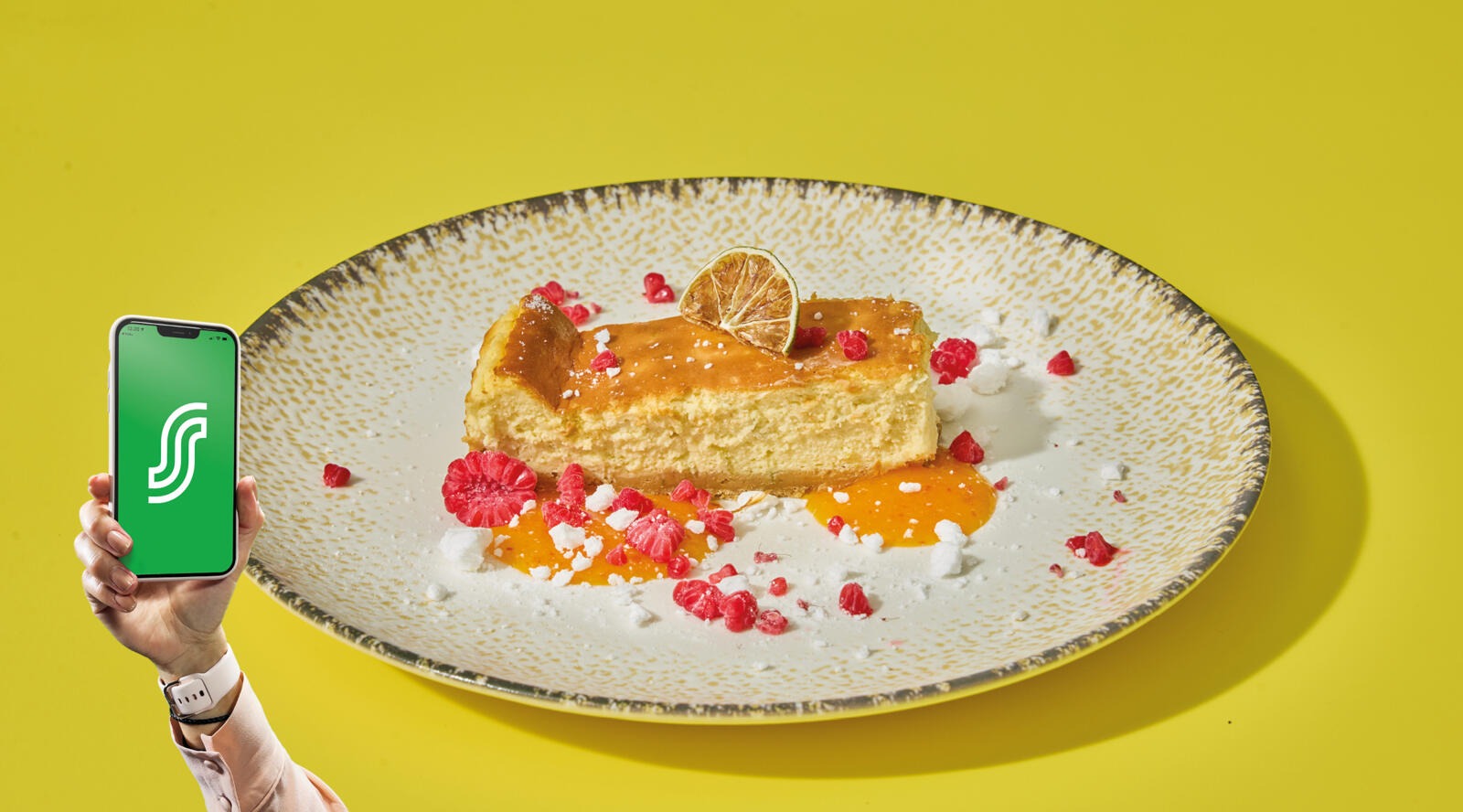 Cheesecake garnished with lime slice and raspberries on a white plate against yellow background. Next to it, a hand holding a smartphone.