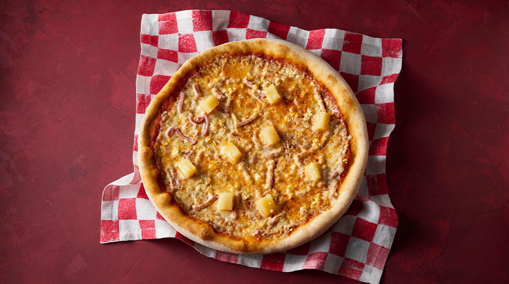 Ham and pineapple pizza on a red and white checkered cloth on a red table, seen from above.