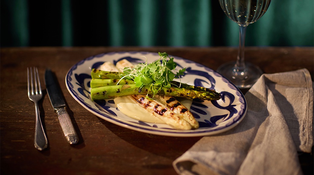 Grillitud kala ja spargel sinise äärega taldrikul, kõrval söögiriistad, salvrätik ja veiniklaas pruunil laual.