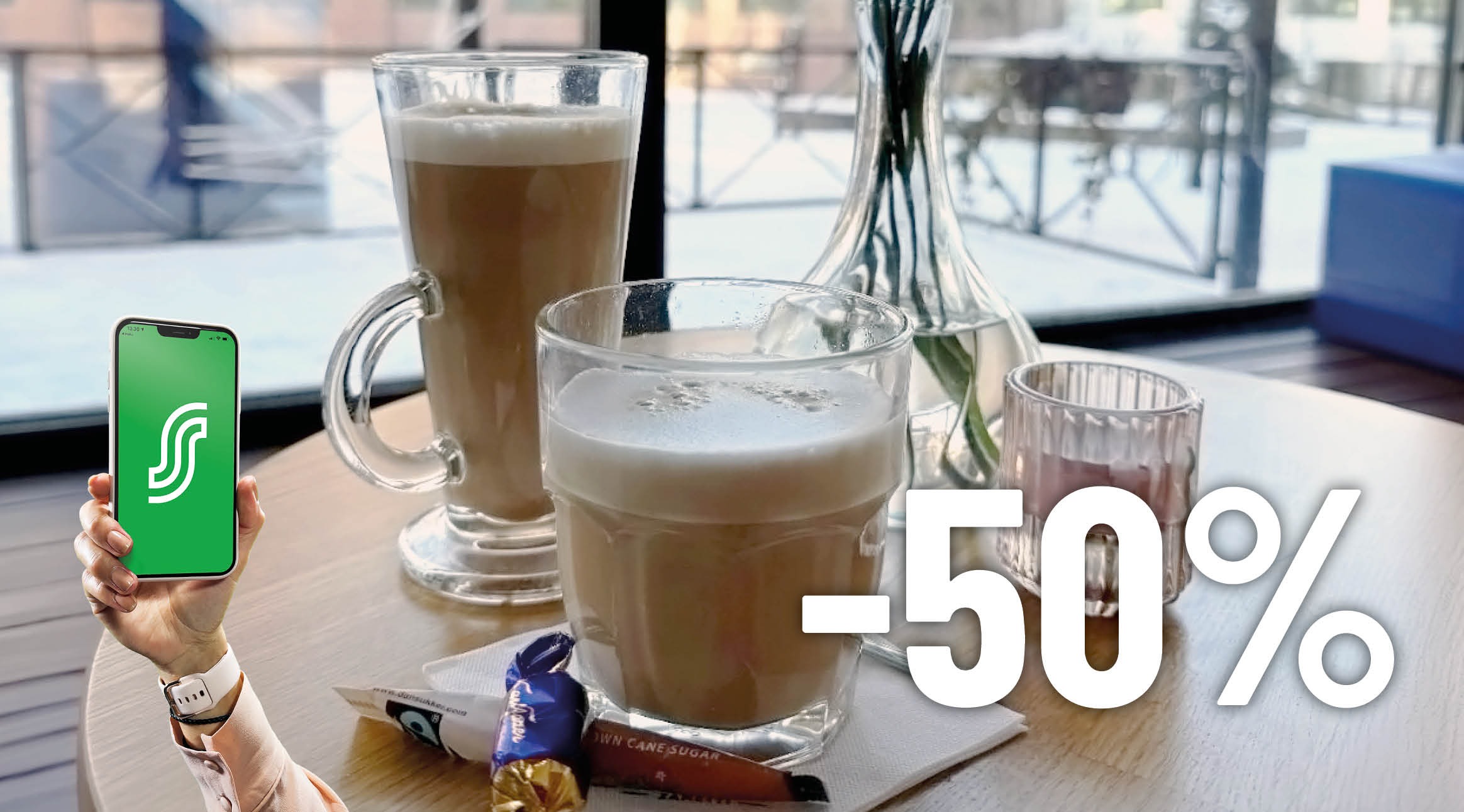 Kahvilapöytä, jolla kaksi latte-juomaa ja käsi pitelemässä älypuhelinta vihreällä sovelluksella. Ikkunasta näkyy talvimaisema