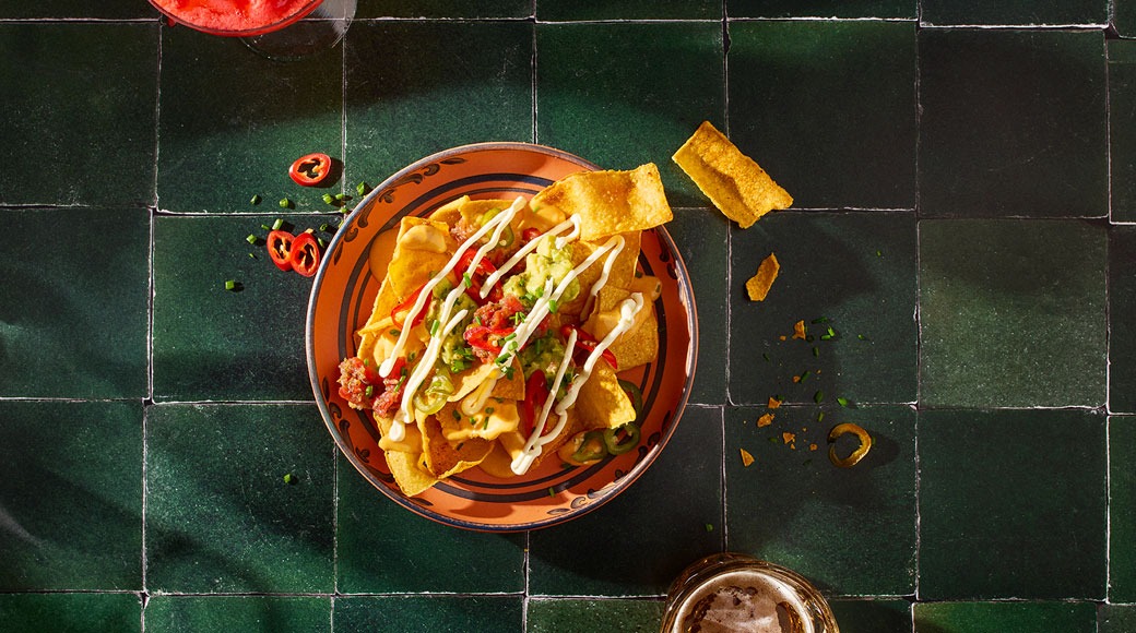 Nachod guacamole, värske tšilli ja kreemja kastmega oranžil taldrikul rohelisel plaatlaual. Kõrval on joogid.