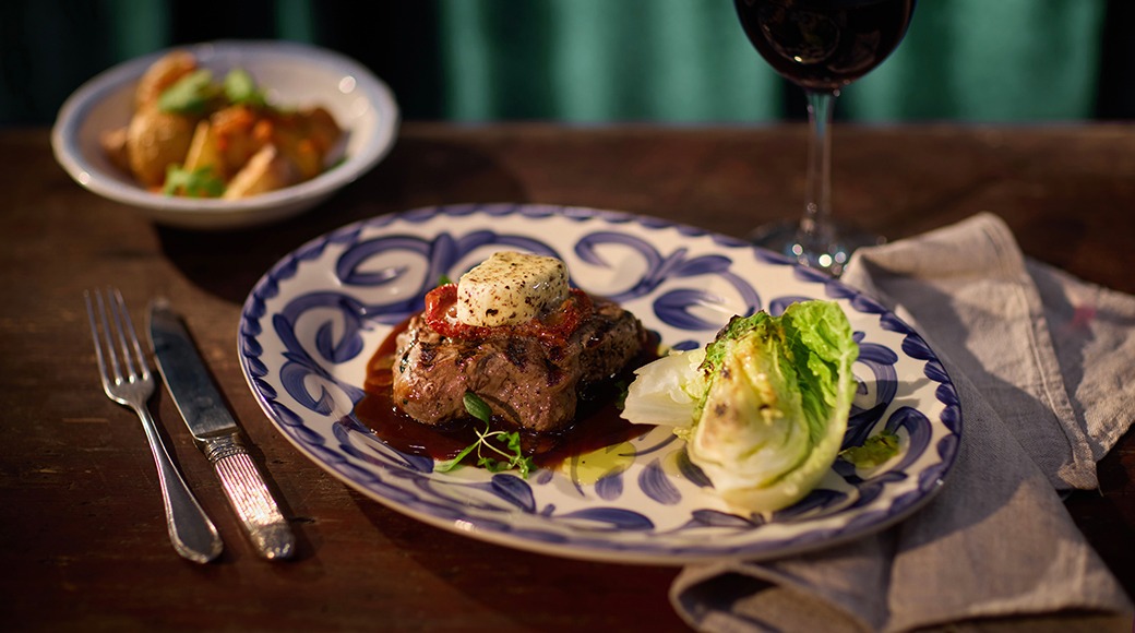 Lihatoit sini-valge kaunistusega taldrikul, peal või, kõrval salat. Taustal veinipokaal ja teine toiduportsjon.