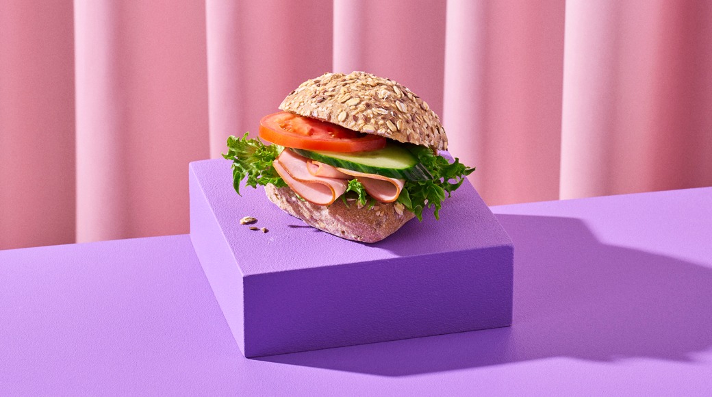 Seesamileipä, jossa kinkkua, tomaattia, kurkkua ja salaattia, violetin korokkeen päällä vaaleanpunaista verhoa vasten.