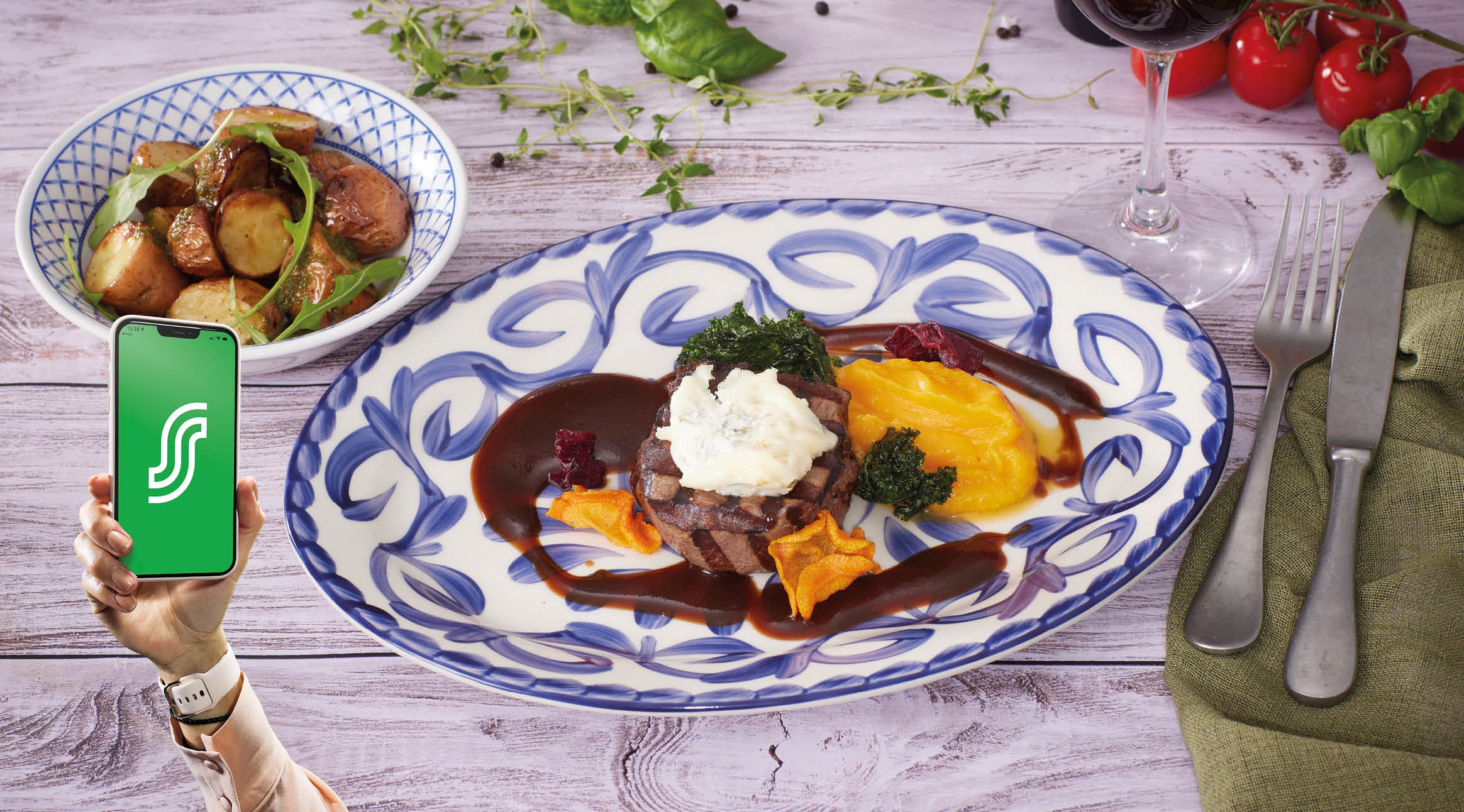 Ateria valkoisella puupöydällä: sinivalkoinen lautanen jossa pihvi ja kastiketta, kulho paistettuja perunoita, viinilasi, käsi pitää älypuhelinta.