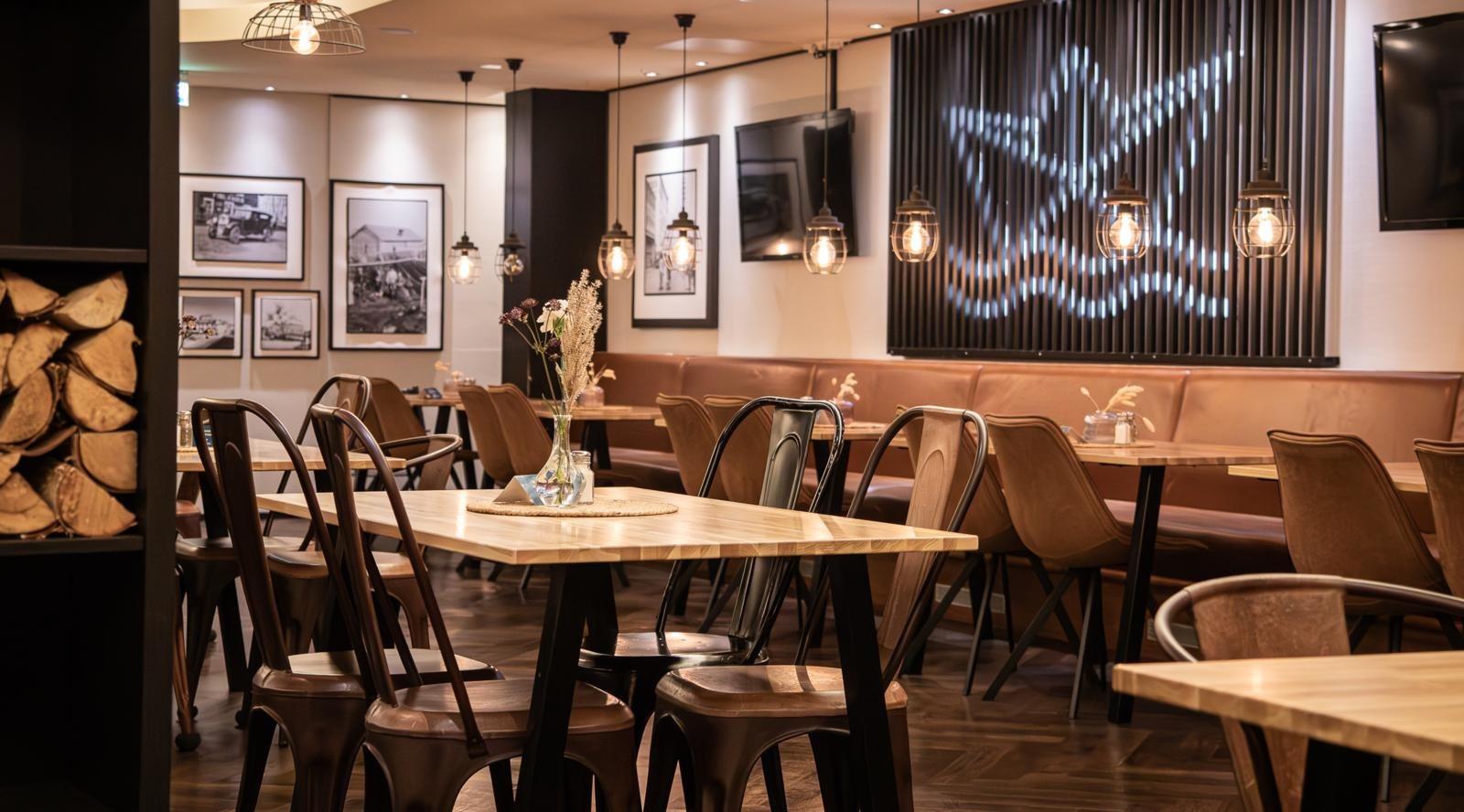 Stylish restaurant with wooden tables and brown chairs. Black and white photographs on walls, pendant lights hanging from ceiling. Small floral arrangement on table.