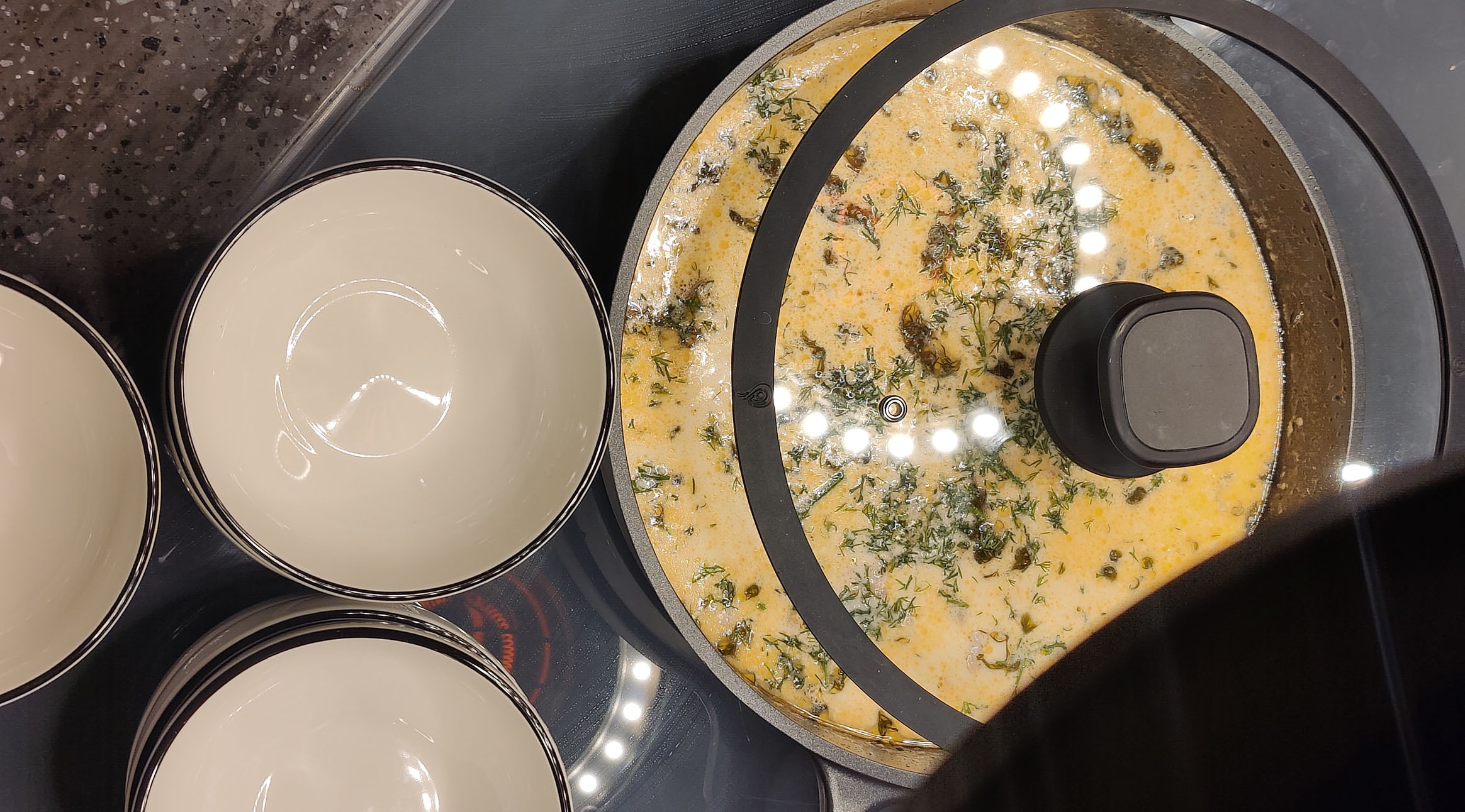 Creamy soup with dill cooking in a pan. Empty ceramic bowls sit beside it on an induction stovetop.