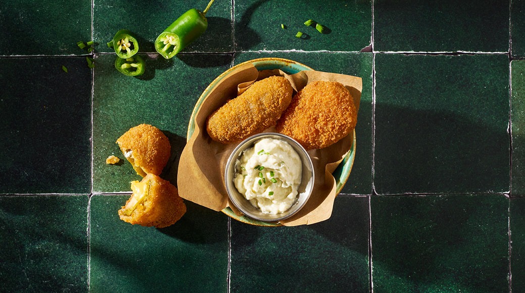 Krõbedad paneeritud jalapeño-juustuampsud paberiga vooderdatud kausis, kõrval kreemjas kaste murulauguga. Tumerohelisel plaadil on pooleks murtud ampsu ja viilutatud jalapeñosid.