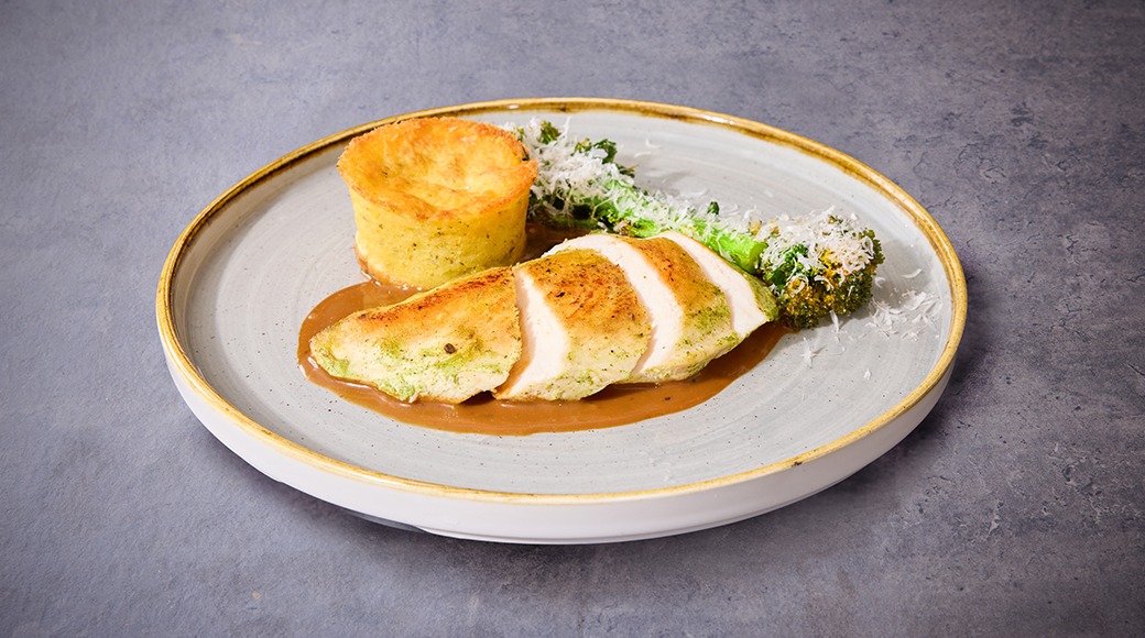 Kana-annos valkoisella lautasella, jossa viipaloitua rintafileetä, pieni sufleemunakas ja parmesan-parsakaalia ruskeassa kastikkeessa.