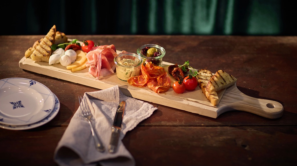 Ett träfat med antipasti: ostar, charkuterier, rostat bröd, oliver och körsbärstomater. Bredvid finns en tallrik och bestick.