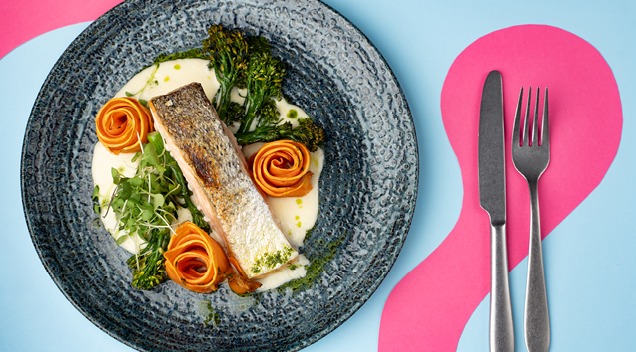 En portion stekt fisk med vit sås, grönsaker och morotsrosor på en blå tallrik, rosa duk.