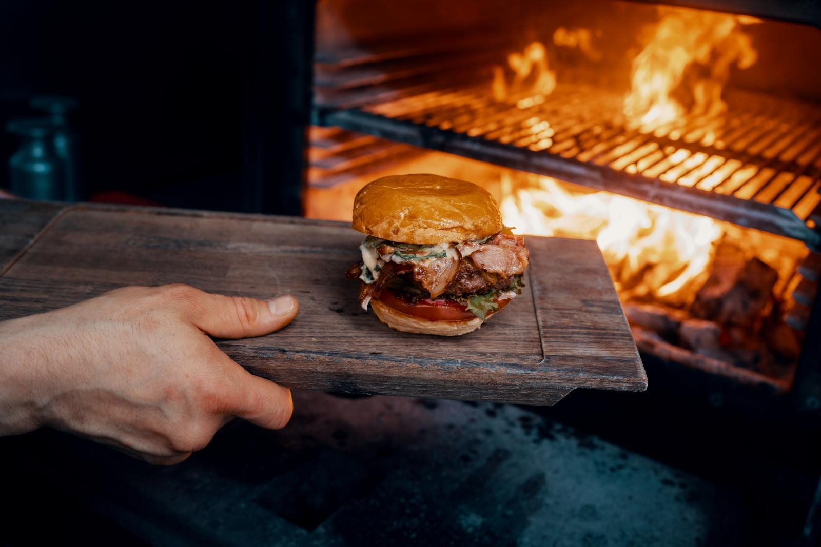 A hand holding a wooden serving board with a loaded burger. An open flame grill blazes in the background.