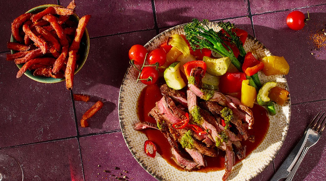 Viipaloitua grillattua lihaa, kastiketta ja yrttitahnaa sekä paahdettuja kasviksia lautasella. Vieressä kulho bataattiperunoita.