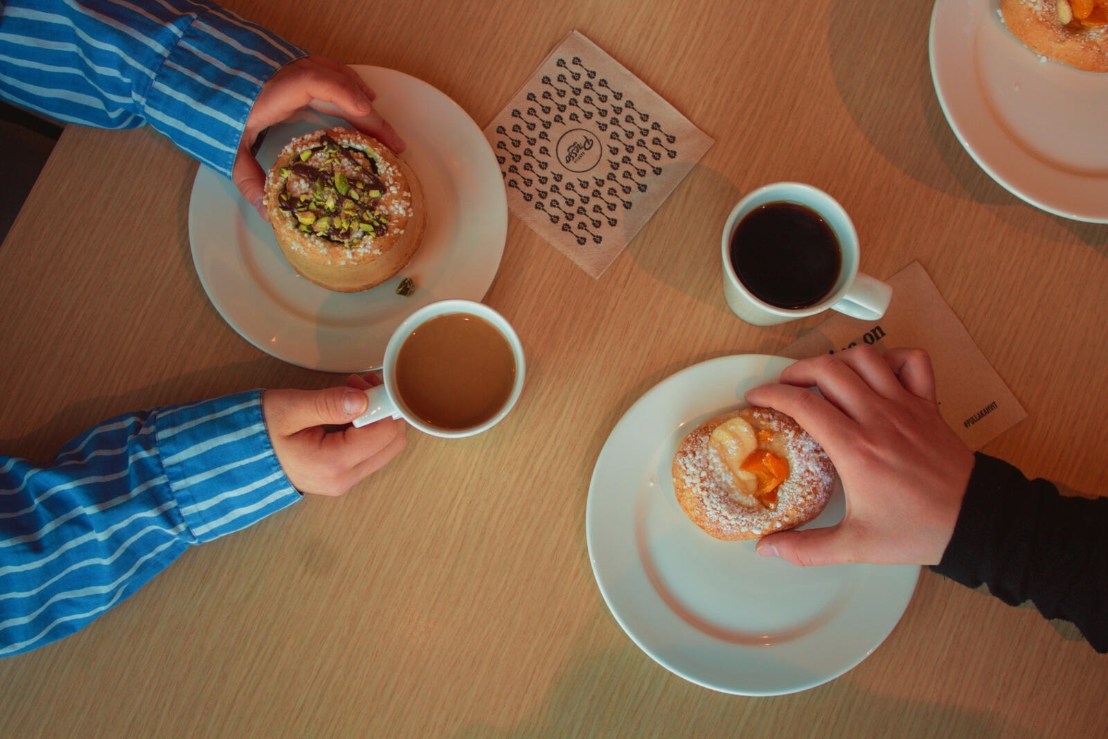 Två personer njuter av kaffe och bakverk vid ett träbord. En äter en pistagebakelse, den andra en aprikosbakelse.