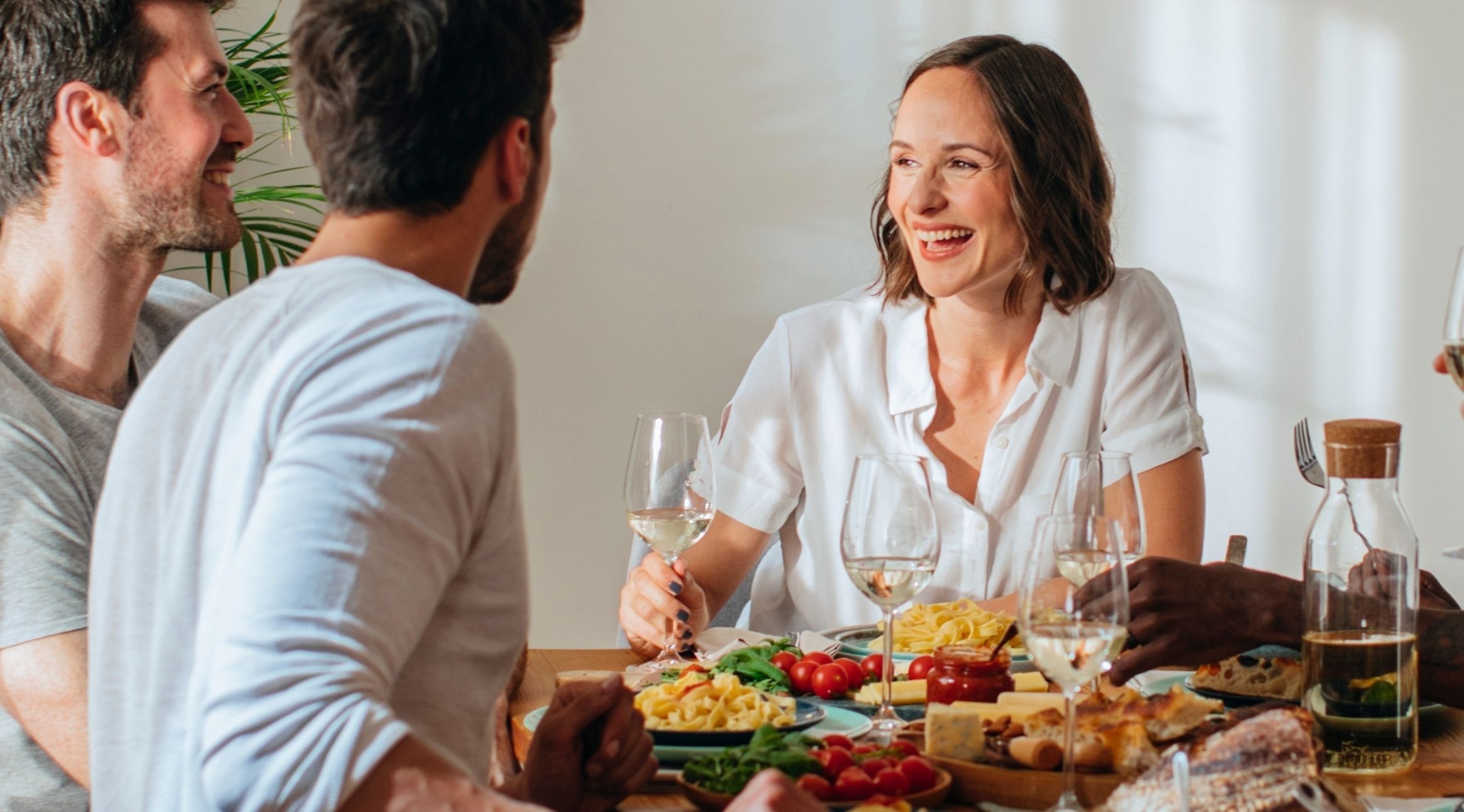 Vänner njuter av en måltid och vitt vin vid ett bord med ostar, grönsaker och pasta.