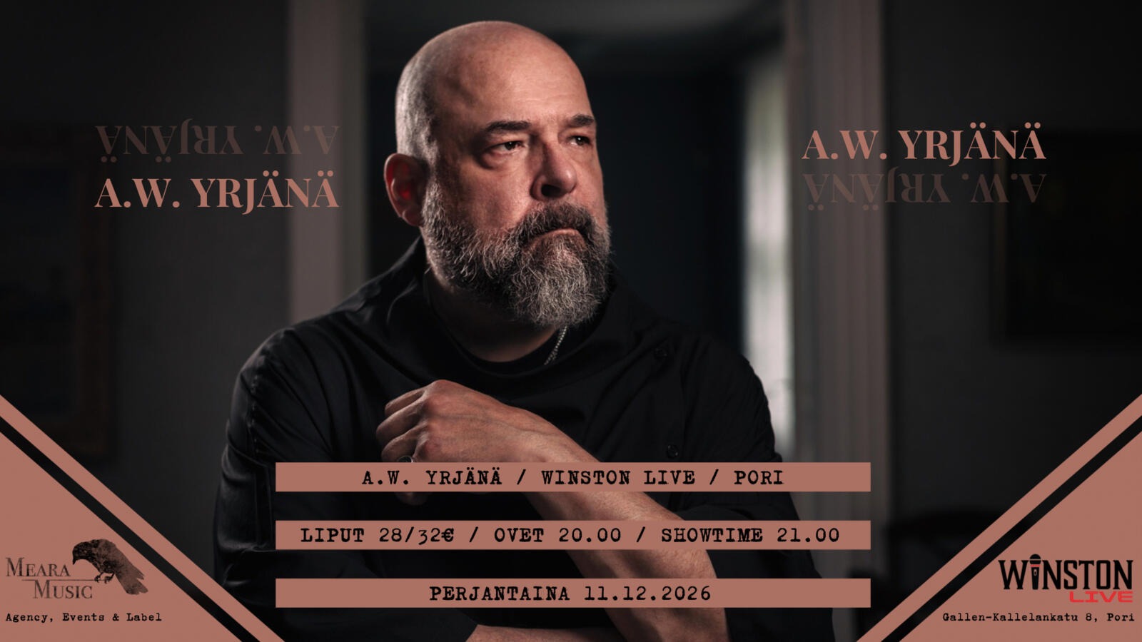 Bald man with beard wearing dark shirt looks directly at camera in dim lighting.