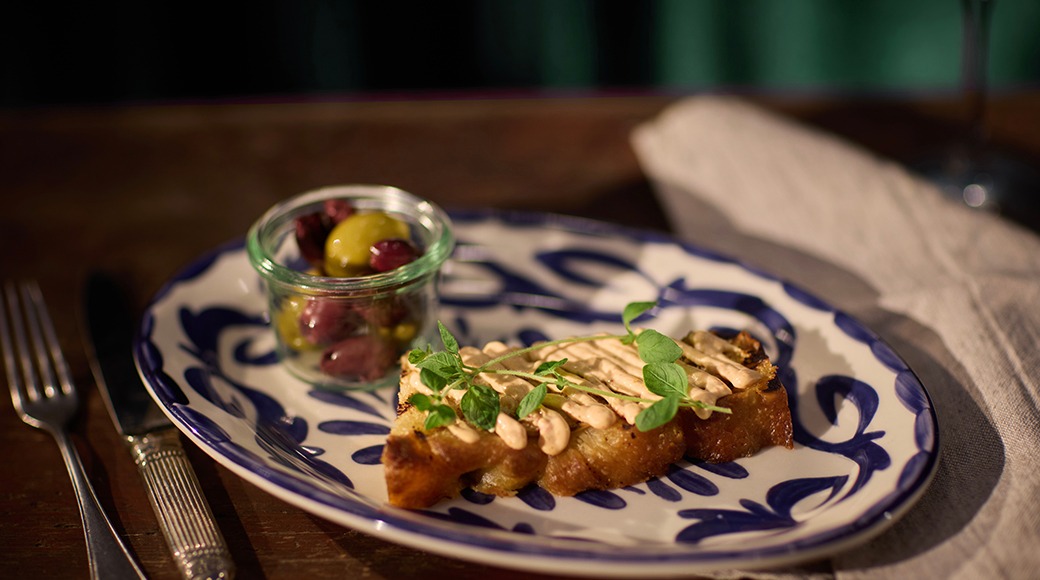 Sinivalge taldrik röstitud leivaga, mida kaunistavad rohelised lehed ja kaste, kõrval väike klaaspurk oliividega.