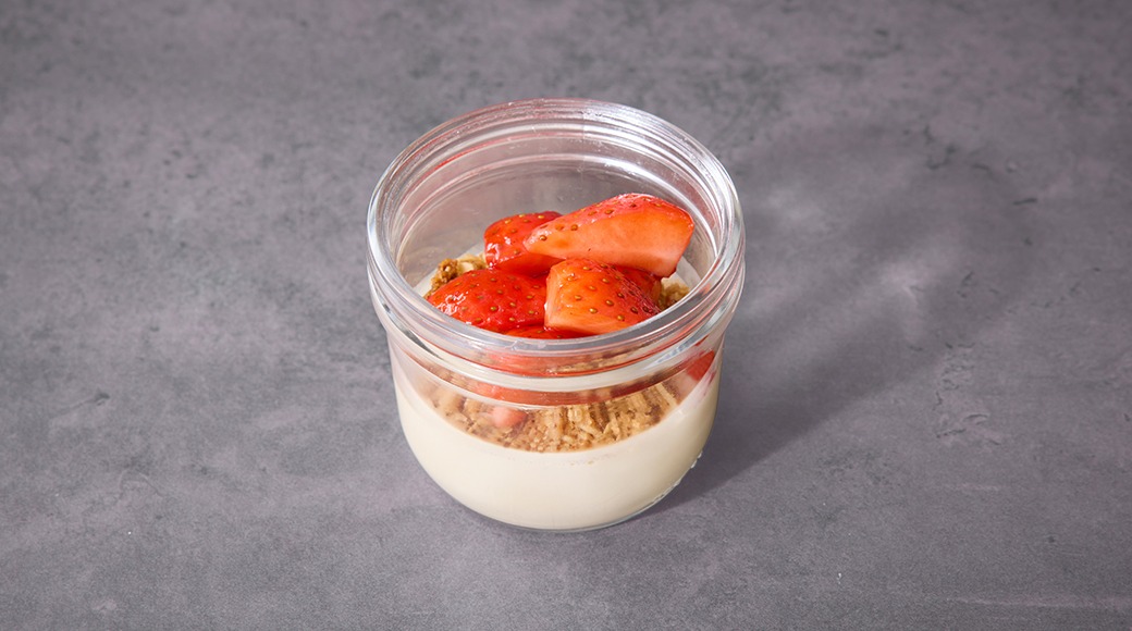 Lasipurkissa oleva jälkiruoka, jossa kerrokset vaniljavanukkaa, murustettuja keksejä ja tuoreita mansikoita.