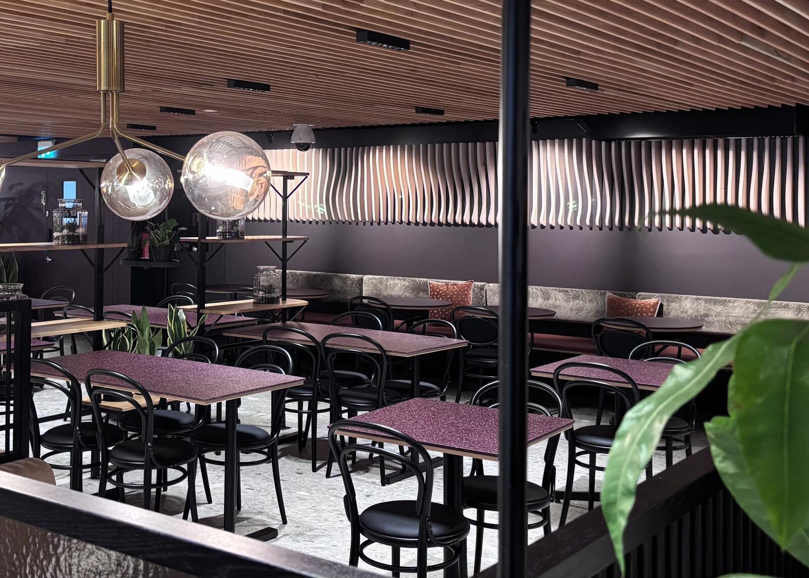 Modern restaurang med violetta bord, svarta stolar och stilfulla klotlampor. Rymlig planlösning, träpaneler i taket och gröna växter.